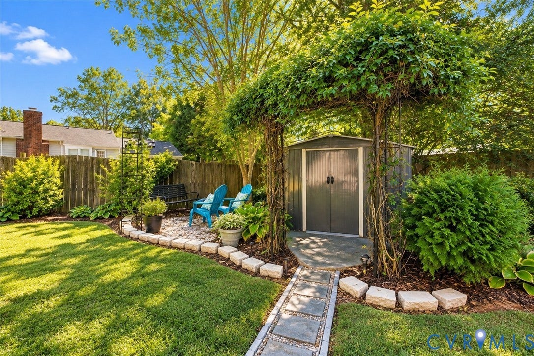 Manicured lawn with a stone-edged garden bed, mature landscaping, and a stepping stone pathway leading to a storage shed with an arched vine-covered trellis