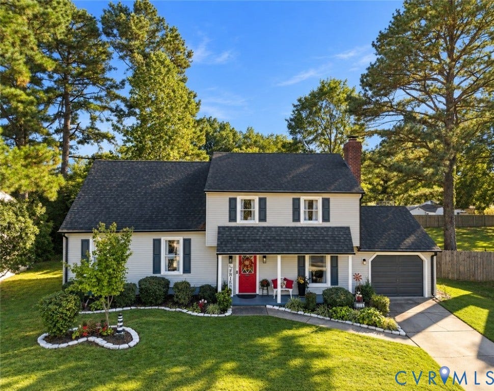 Two-story property featuring light-colored siding, a covered entry porch, an attached garage, a brick chimney, and mature trees surrounding the property