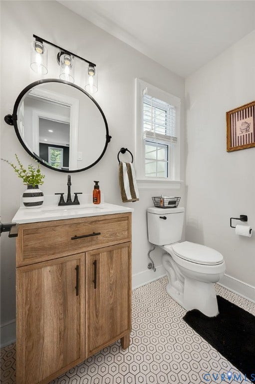 13800 W Salisbury Road, Salisbury, Midlothian, VA 23113 Wood-finish vanity with a white countertop and integrated sink, matte black faucet, oval mirror, and hexagonal floor tiling