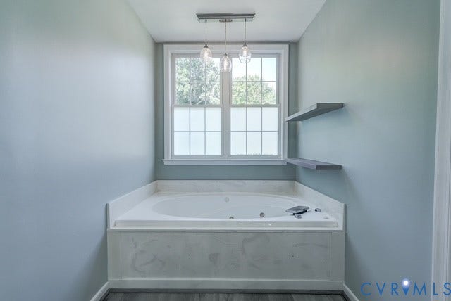 8113 Ainsdale Lane, Chesterfield, VA 23832 Soaking tub with a white surround and integrated faucet, under a window with privacy film, illuminated by a multi-pendant light fixture, complemented by two floating shelves