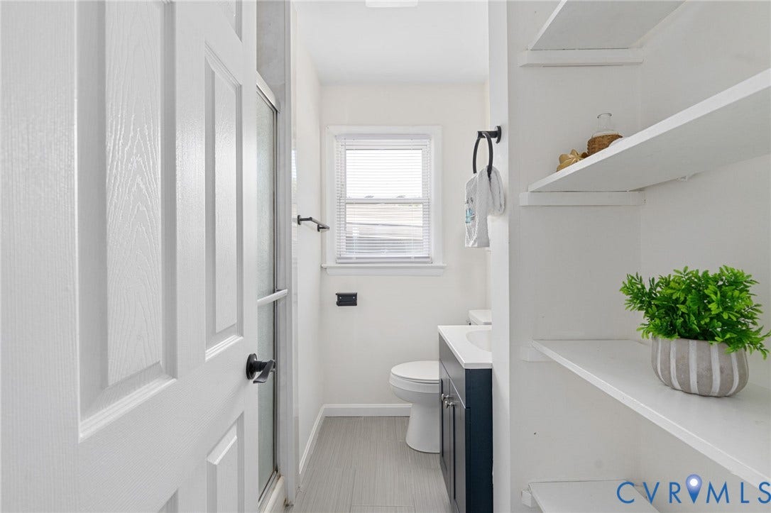 Built-in shelving, white paneled door, shower enclosure with obscure glass, single vanity with dark cabinetry, and a window with blinds