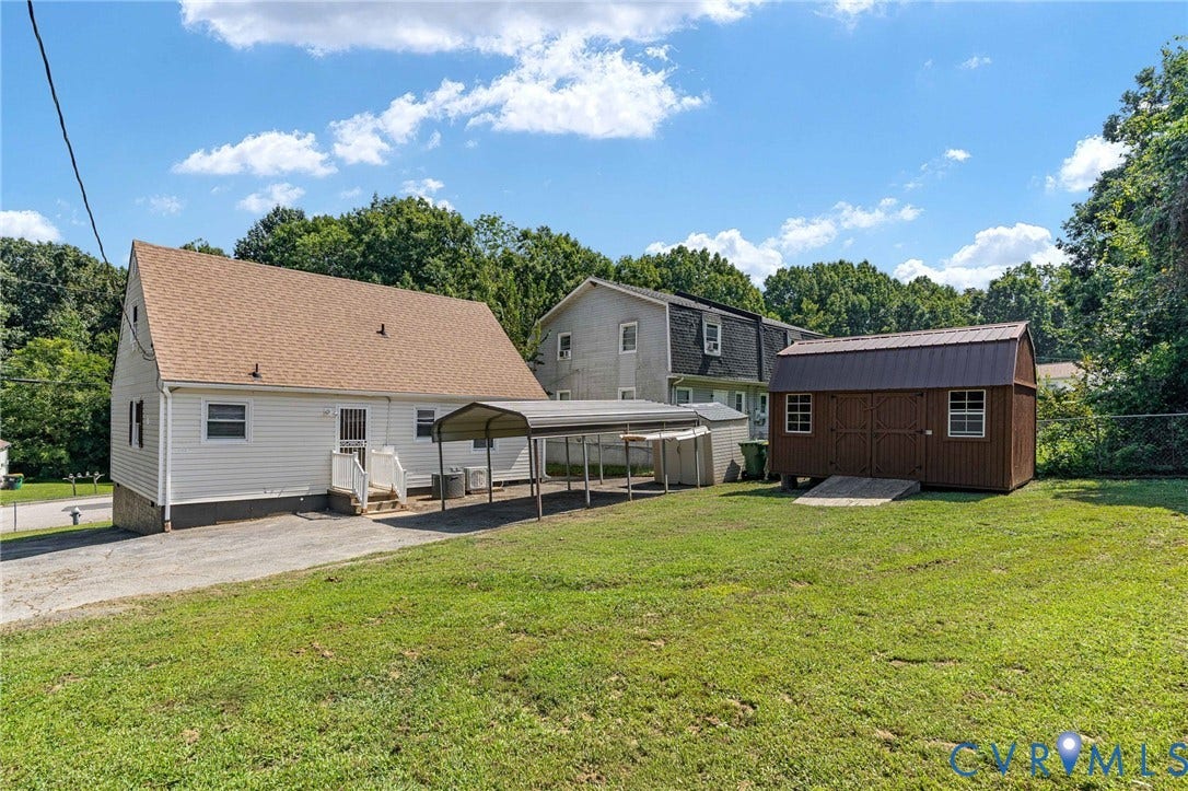 Expansive grassy yard featuring a brown storage shed, gravel driveway, and a covered carport