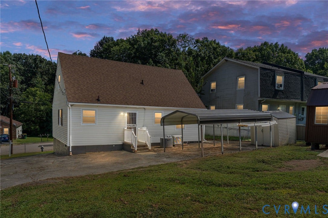 White siding exterior with a brown shingle roof, rear entry with white railing, and a detached metal carport