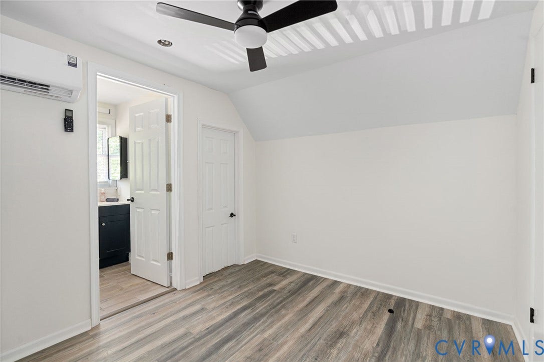 Sloped ceiling room featuring wood-finish flooring, a ceiling fan, a wall-mounted climate control unit, and a doorway to an adjoining space with a dark vanity