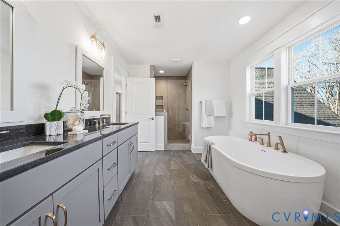 9169 Terroir Lane, Villas, New Kent, VA 23124 Freestanding soaking tub with brass fixtures, dual vanity with undermount sinks and dark countertops, walk-in shower with tiled walls, and large windows