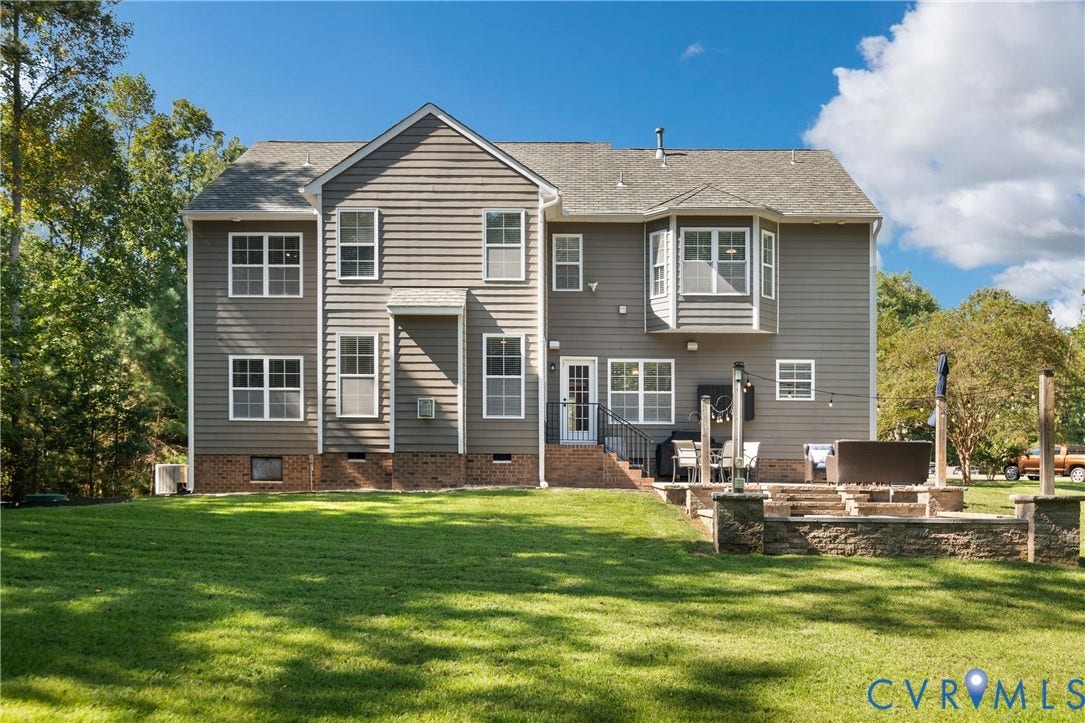 Rear facade with gray horizontal siding, bay window, white-framed windows, brick foundation, and multi-level stone hardscaping