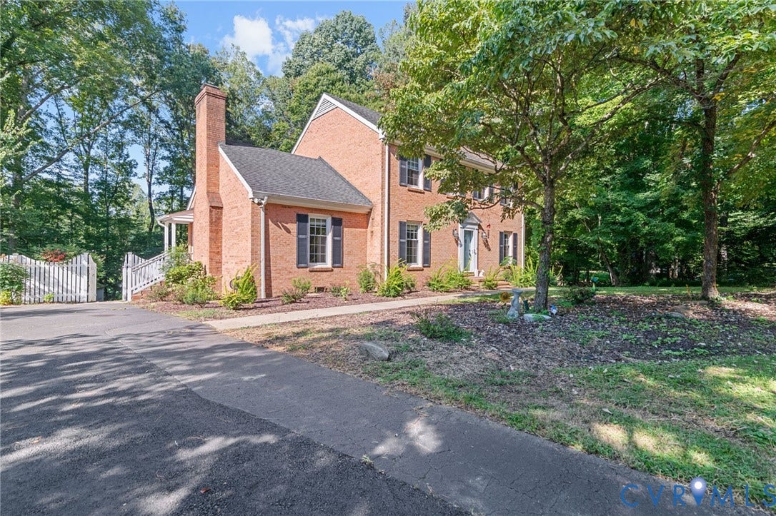 Brick exterior with black shutters, mature trees, a paved driveway, and a white picket fence