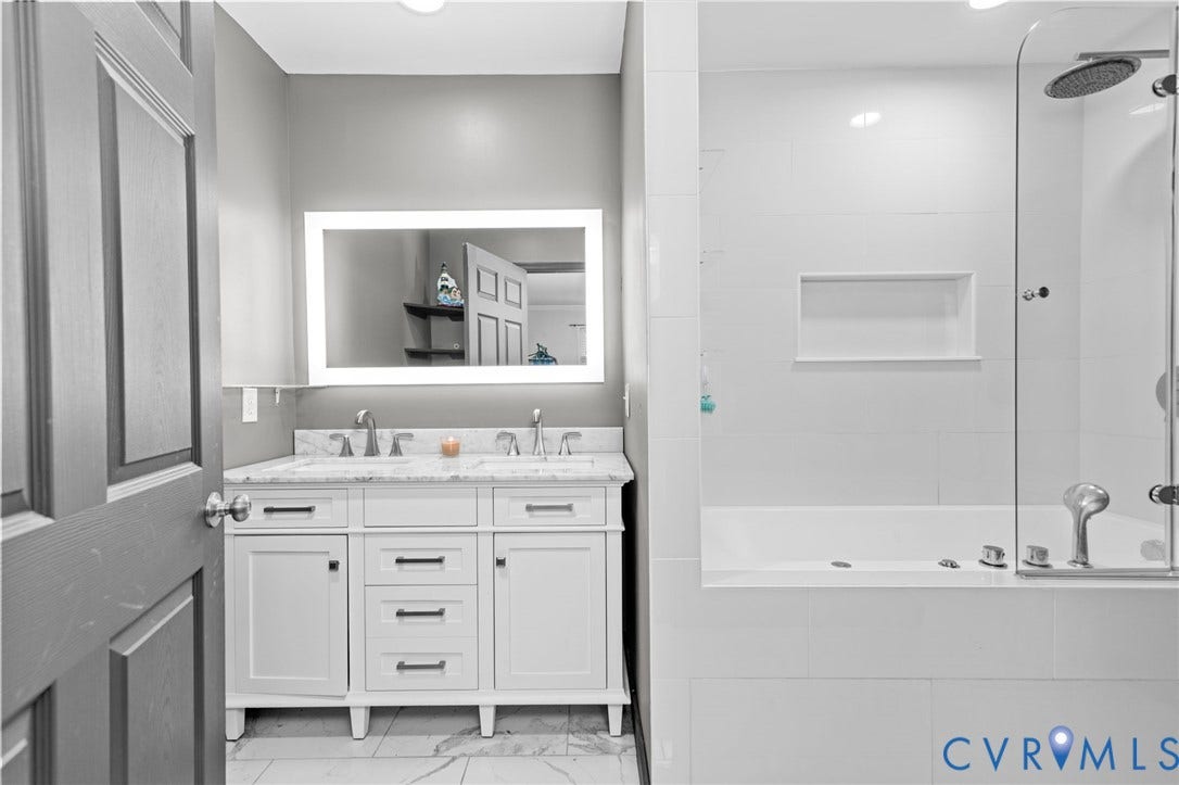 Dual vanity with stone countertop and undermount sinks, illuminated mirror, and tiled shower/tub combination with glass enclosure