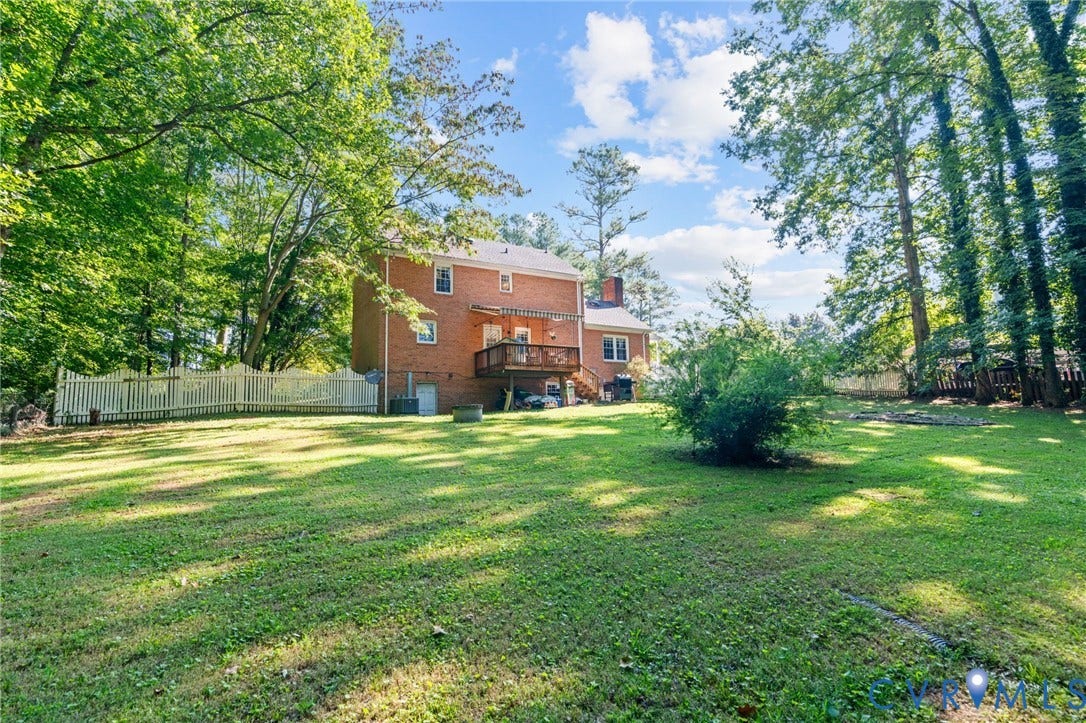 Expansive green lawn with mature trees, a white picket fence, and a brick exterior home with an elevated wooden deck