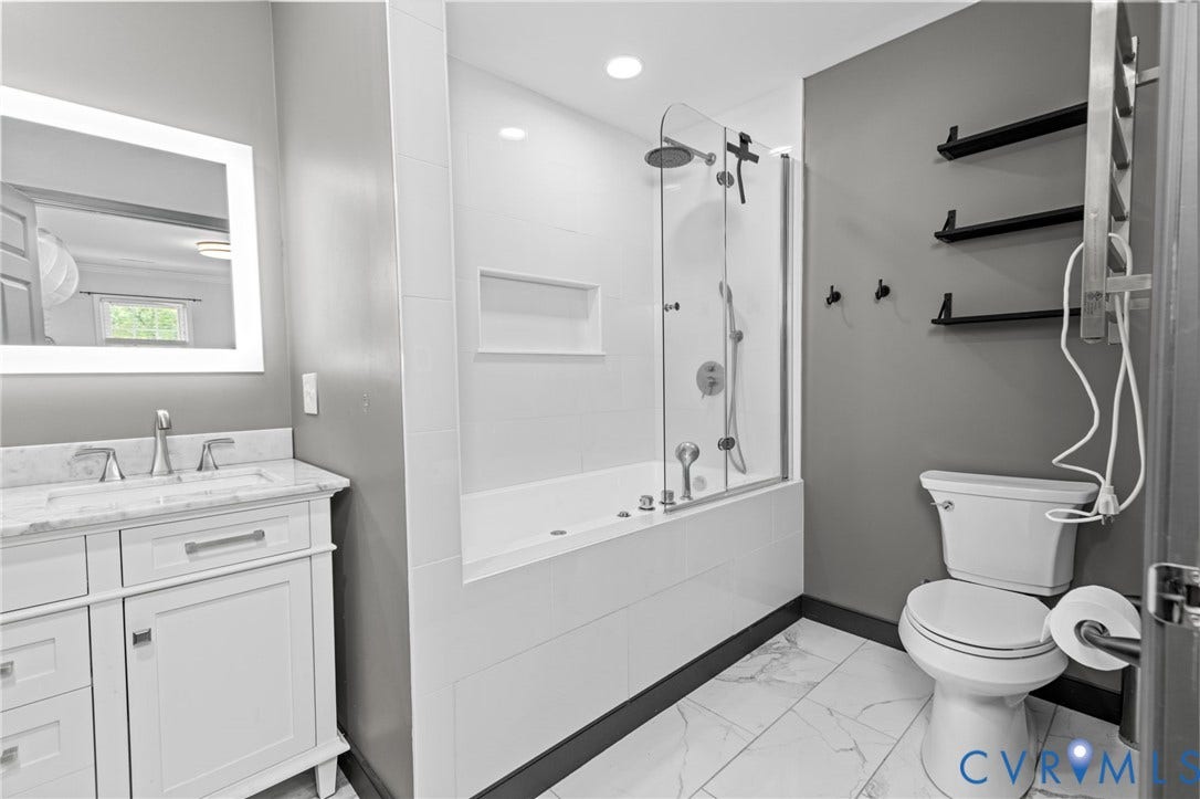 Modern bathroom featuring a white vanity with a marble-look countertop, an illuminated mirror, a built-in tub with a glass enclosure, and large format floor tiles
