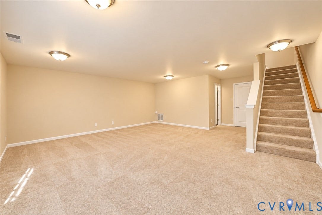 Expansive carpeted area with neutral-toned walls, multiple ceiling-mounted light fixtures, and a carpeted staircase with a wood-finish handrail