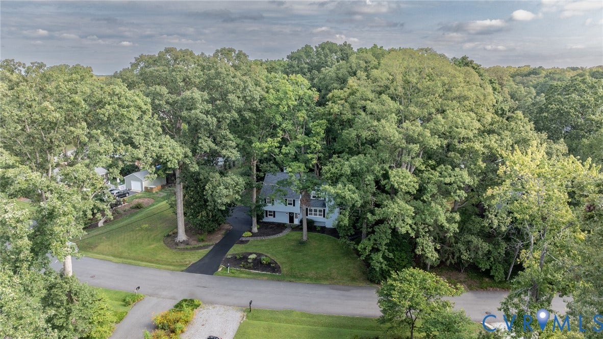 Paved driveway leading to a two-story residence with light siding and dark shutters, surrounded by extensive mature trees