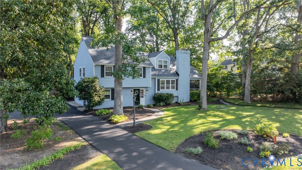 Light blue siding exterior with black shutters, dormer windows, a prominent chimney, an asphalt driveway, and mature trees