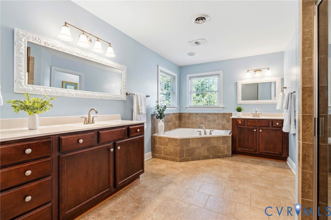 Dual vanity countertops, rich wood-finish cabinetry, an inset soaking tub, tiled tub surround, and a walk-in shower with tiled walls