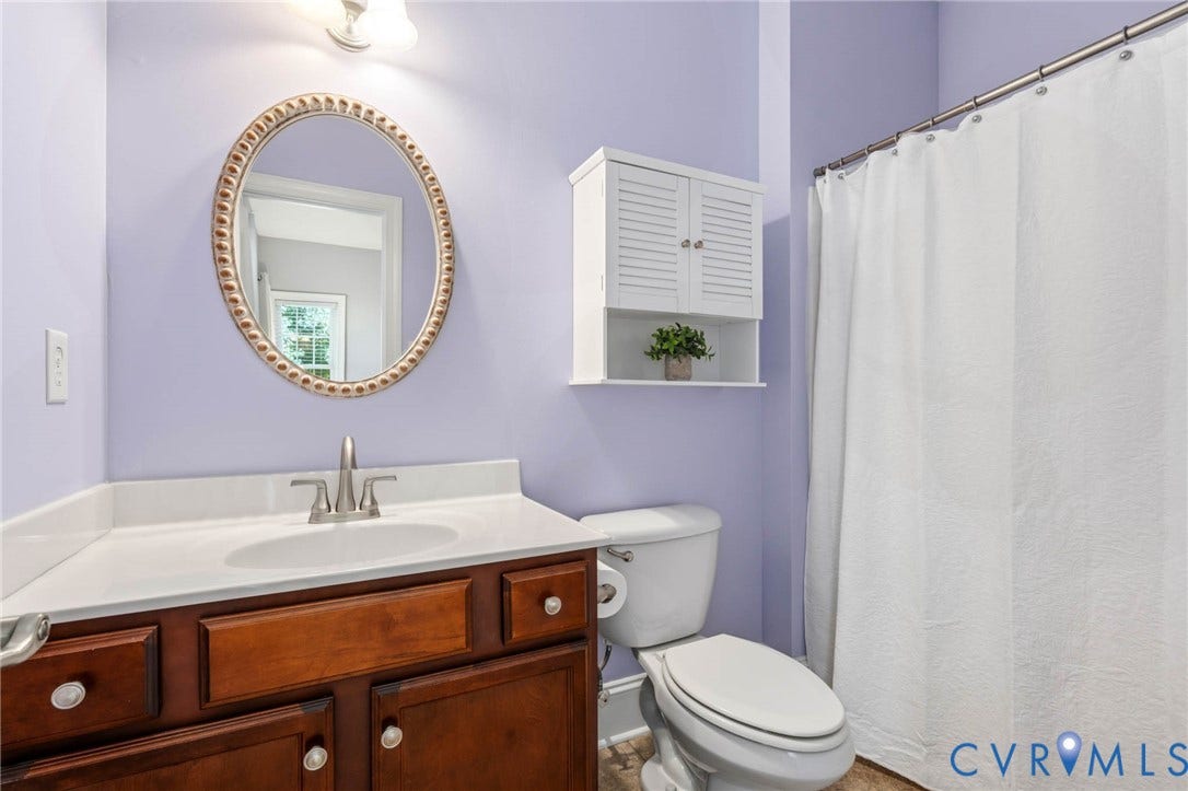 First Floor Ensuite Bathroom featuring a wood-finish vanity with a white countertop and integrated sink, brushed nickel faucet, an oval mirror, and a wall-mounted storage cabinet