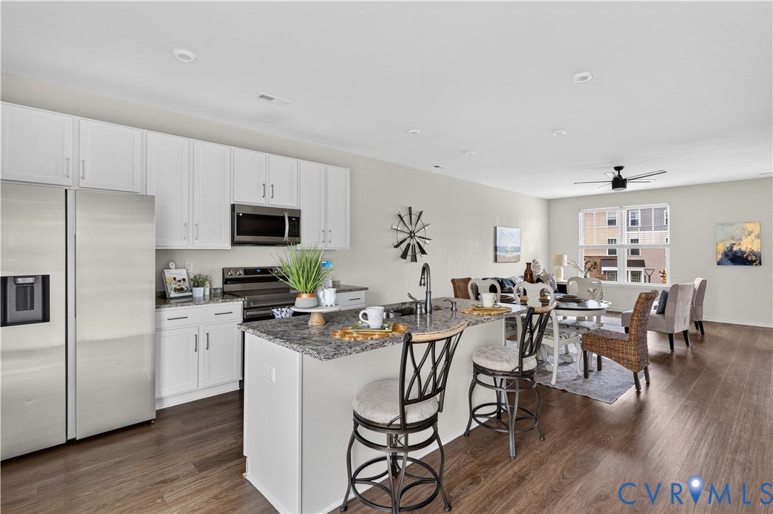 Open-concept layout featuring wood-finish flooring throughout, a kitchen island with stone-finish countertop, white cabinetry, and stainless steel appliances
