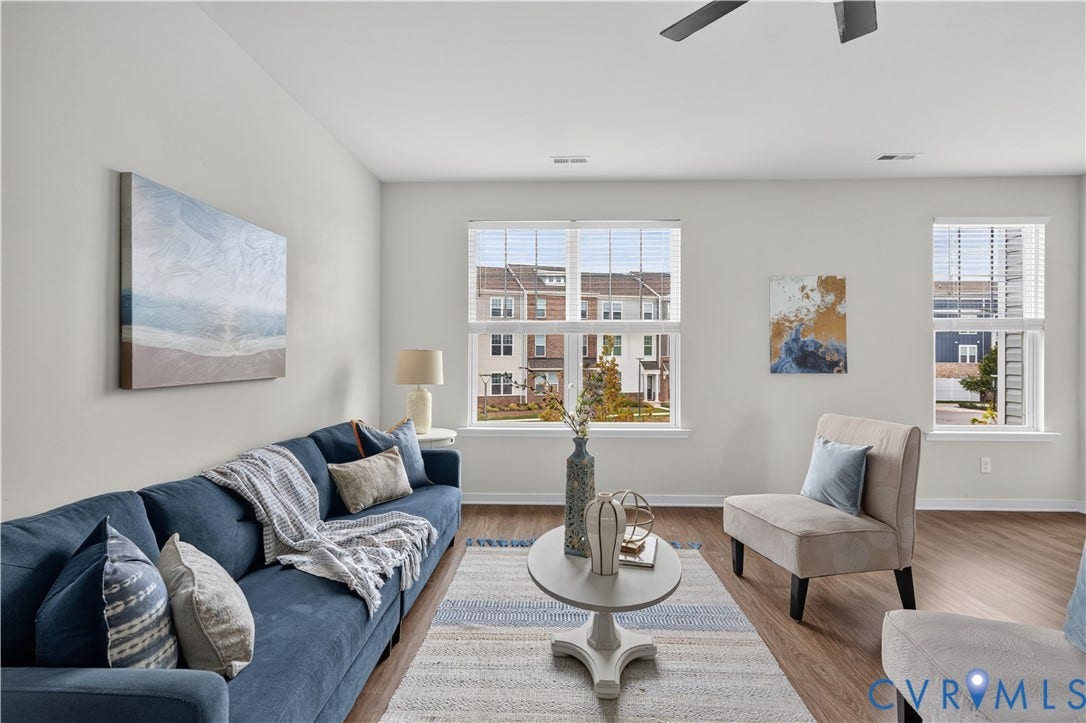 Spacious living area featuring light-toned walls, wood-finish flooring, multiple windows with blinds, and a ceiling fan