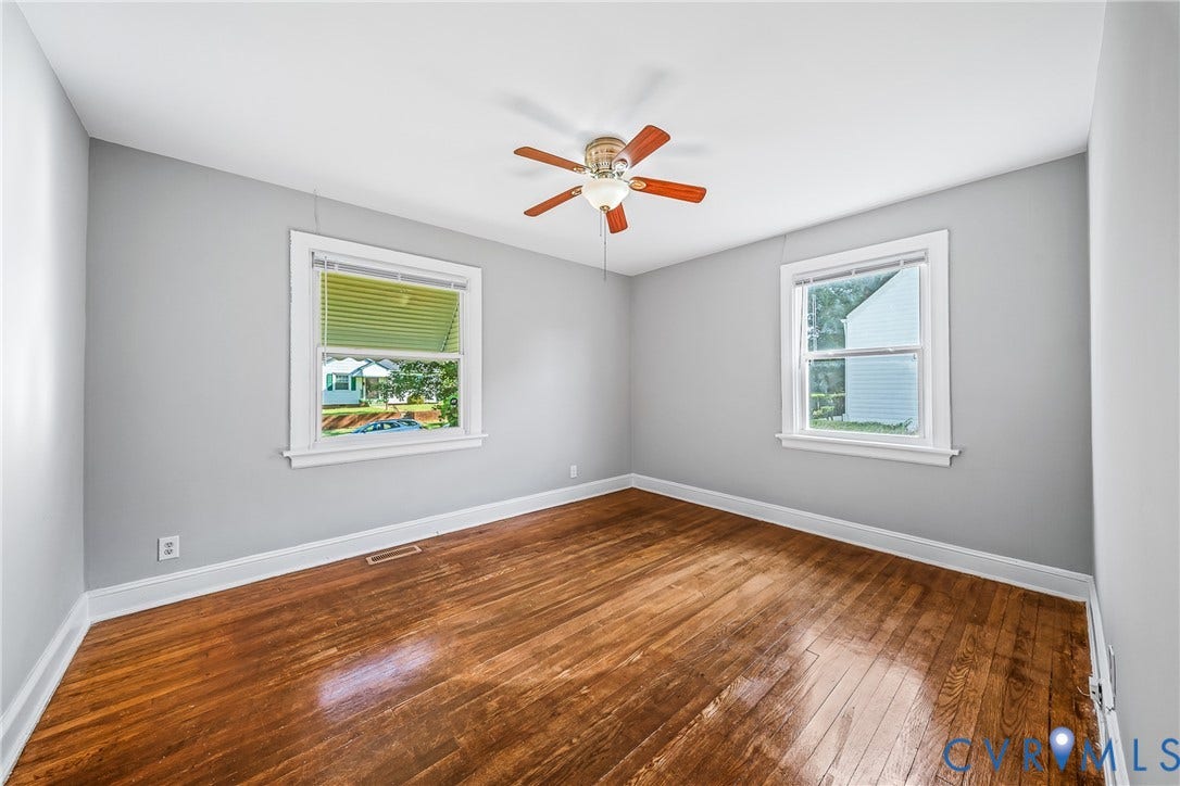 Wood flooring, light gray wall paint, two windows with white trim, and a ceiling fan with wood-tone blades