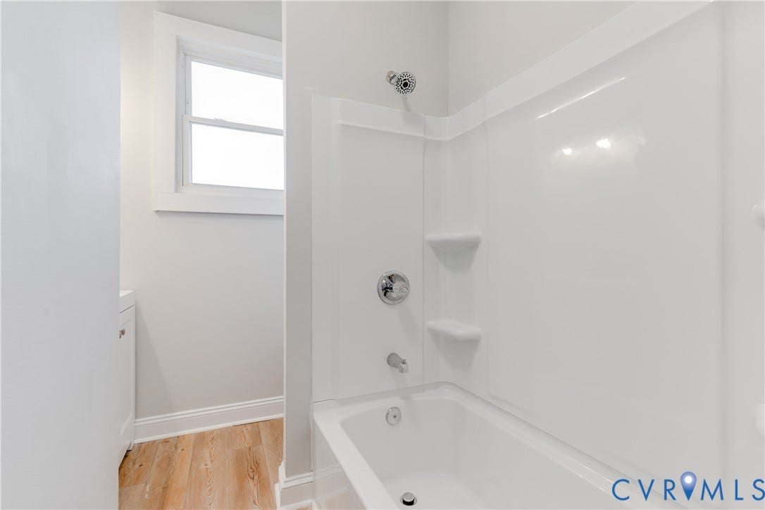 White shower/tub combination with chrome fixtures, integrated shelving, window, and wood-finish flooring