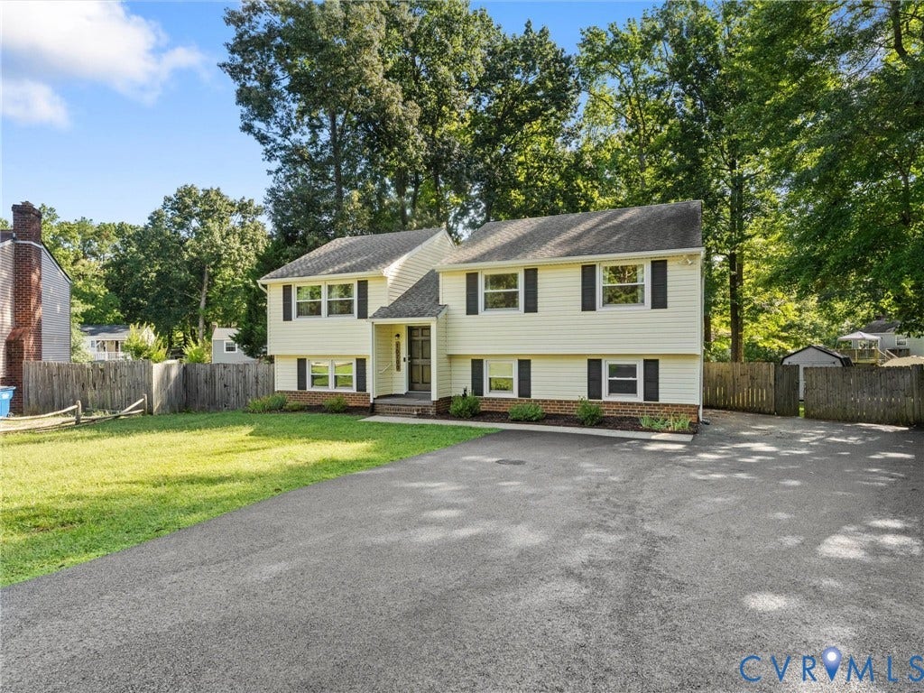 Split-level home featuring light-colored siding, black window shutters, a front entry with a storm door, and a spacious paved driveway