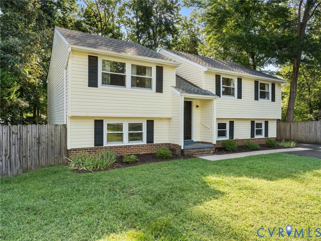 Split-level residence featuring light-toned siding, dark shutters, a covered entryway, mature trees, and a wooden privacy fence