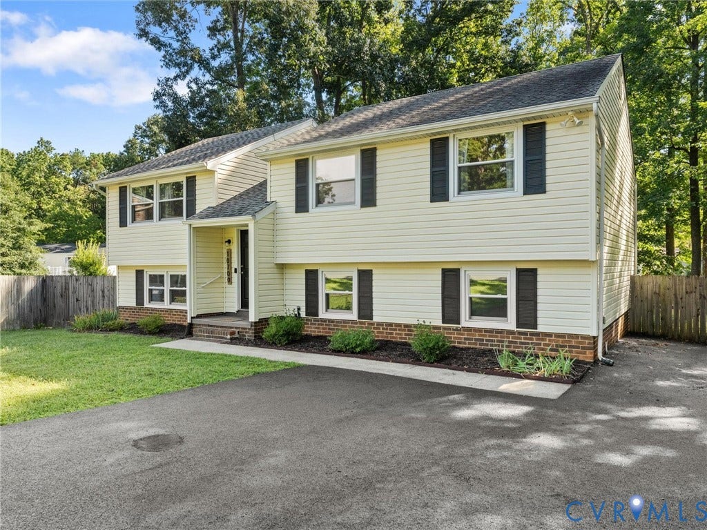Split-level residence featuring light-colored siding, black window shutters, a covered entry, established landscaping, and a paved driveway