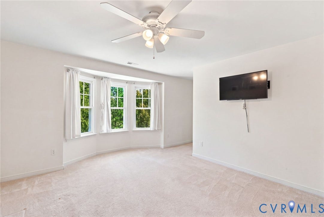 Bay window with multiple panes, ceiling fan light fixture, wall-mounted television bracket, neutral carpeted flooring, and white walls
