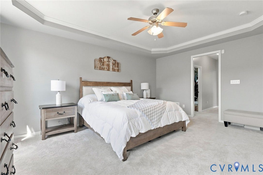 Recessed ceiling with crown molding, ceiling fan, neutral wall paint, light-colored carpeting, and a doorway leading to an additional space