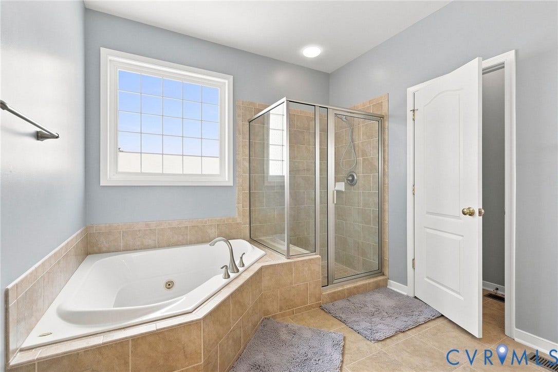 Corner soaking tub with deck-mounted faucet, separate glass-enclosed shower, tiled flooring, and a large window with grid panes