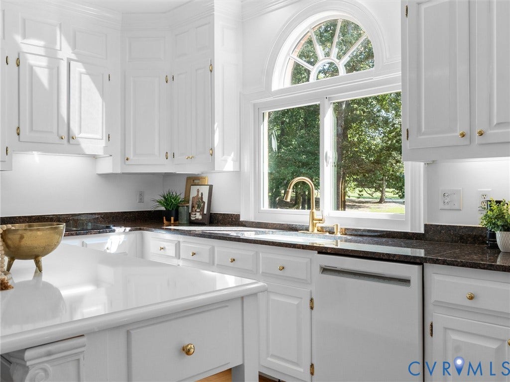 White cabinetry with gold-tone hardware, dark countertops, white dishwasher, and a window with an arched transom