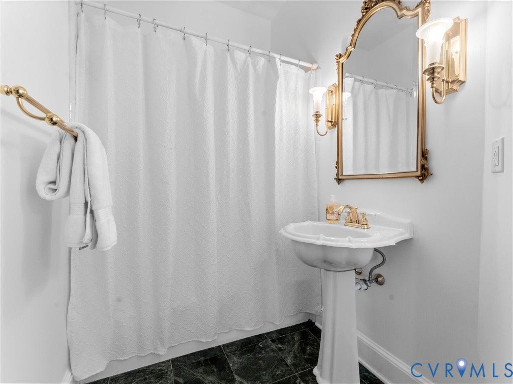 1st floor full bath w/ pedestal sink with gold-finish fixtures, ornate gold-frame mirror, dual wall-mounted sconces, and dark stone tile flooring