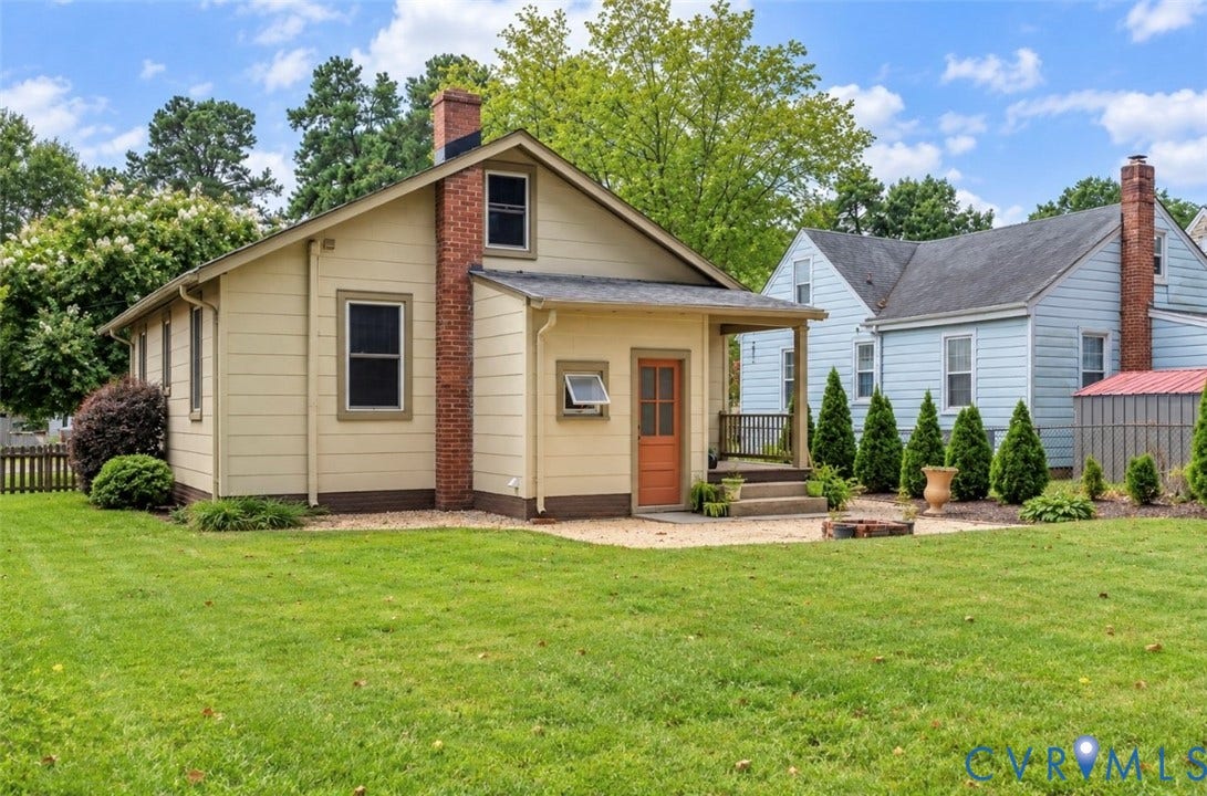lawn area with a gravel patio, covered porch entry, brick chimney, and established perimeter plantings