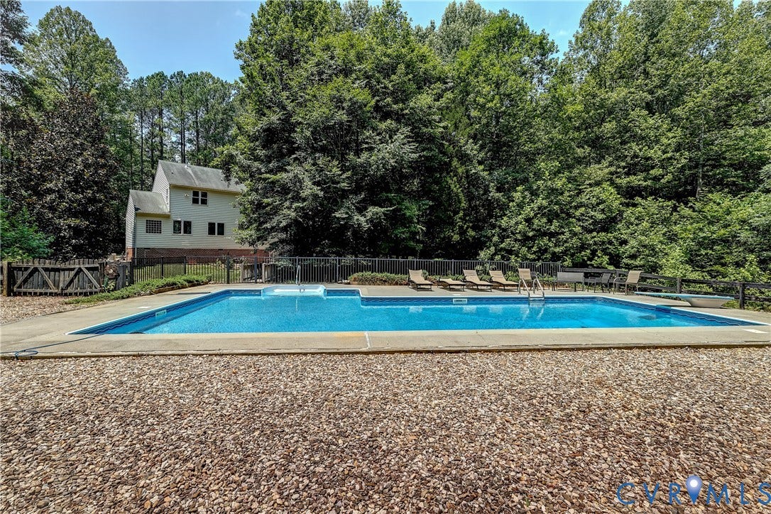 Expansive in-ground swimming pool with surrounding concrete decking, secured by perimeter fencing, set against a backdrop of mature trees