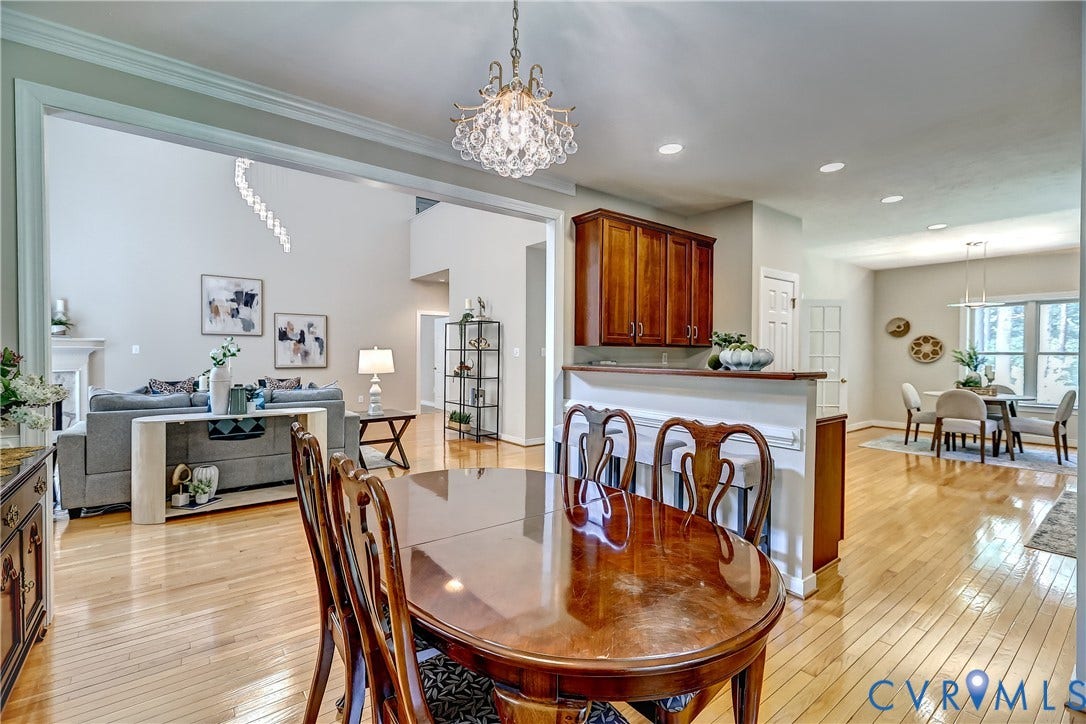 Open-concept dining and kitchen area showcasing hardwood flooring throughout, a round wood-finish dining table with seating for six, custom wood cabinetry with a built-in bar featuring white subway tile backsplash, and a crystal chandelier overhead