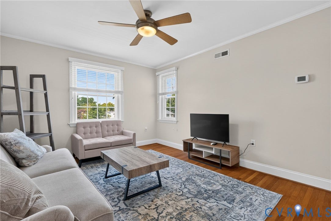 Bright and airy living room showcasing hardwood flooring, large windows with natural light, a ceiling fan with integrated lighting, and a wood-finish coffee table