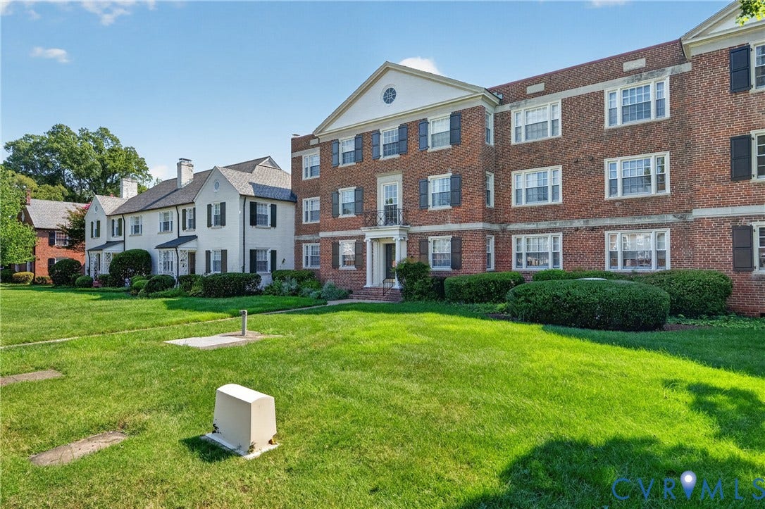 Brick apartment building featuring a gabled portico entrance, multiple windows with dark shutters, and a manicured lawn