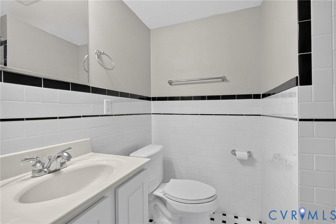 White subway tile with black accent trim, white vanity with integrated sink, chrome faucet, wall-mounted mirror, and black and white floor tiling