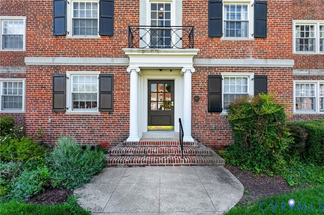 Brick exterior featuring decorative shutters, a columned entryway, a wrought iron balcony, and established landscaping