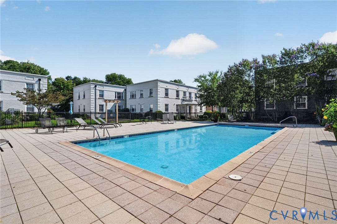 Outdoor swimming pool with a surrounding paved deck, enclosed by a black metal fence, and bordered by mature trees