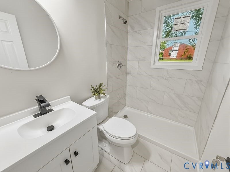 White vanity with integrated sink and matte black faucet, round wall mirror, tiled shower surround with chrome fixtures, and a window with white trim