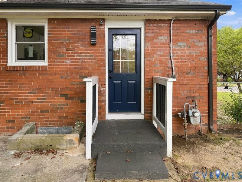 Brick exterior with a dark-colored entry door featuring glass lites, a wall-mounted light fixture, and white railings