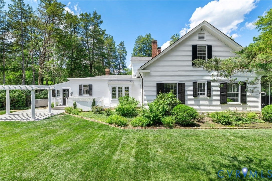 White clapboard exterior with black shutters, a gabled roofline, a patio with a white pergola, and landscaped garden beds