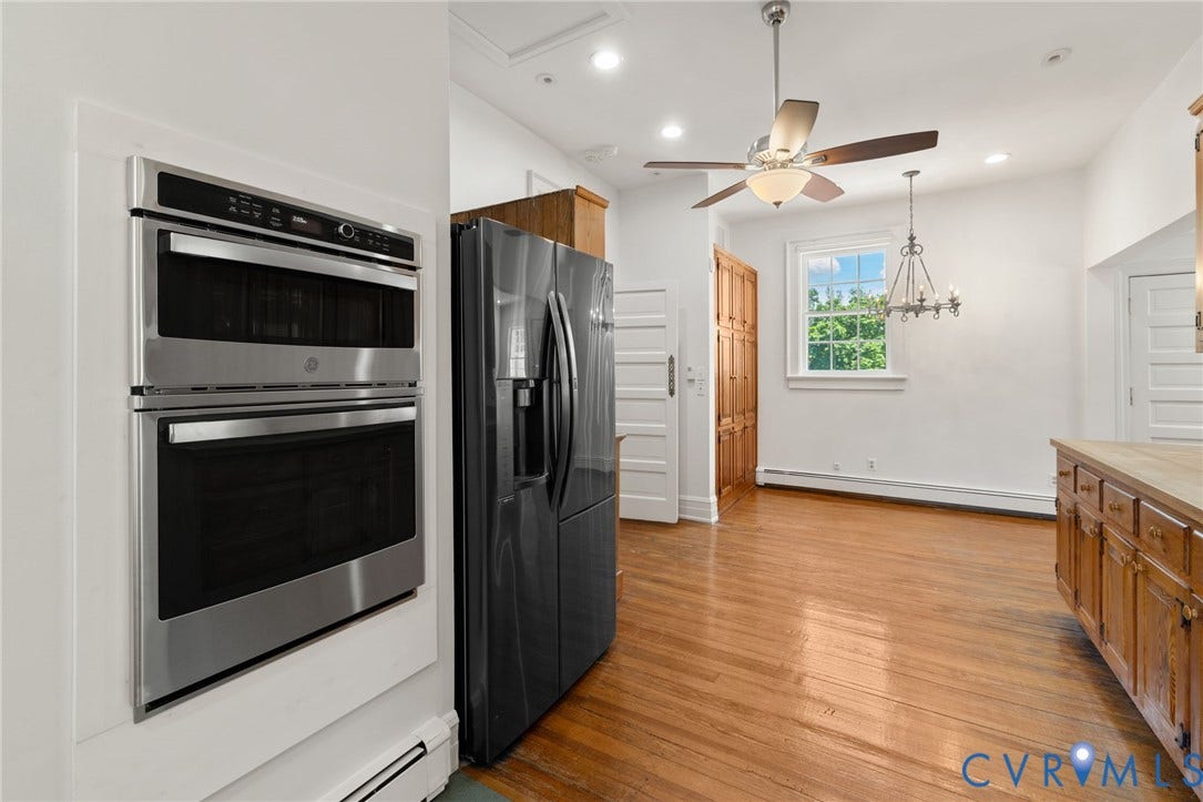 Stainless steel double wall ovens, built-in refrigerator, wood-finish flooring, wood cabinetry with a light-tone countertop, and a ceiling fan with integrated lighting