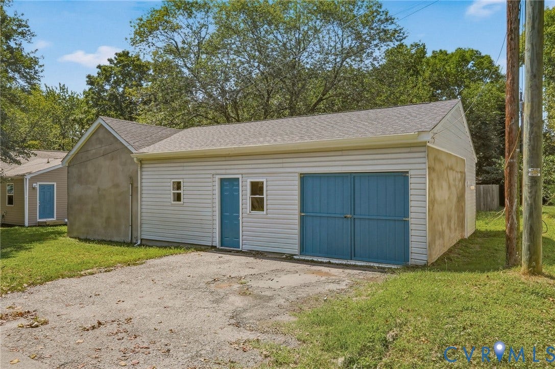Detached garage featuring light-colored siding, a blue pedestrian door, and a blue garage door, with a gravel driveway