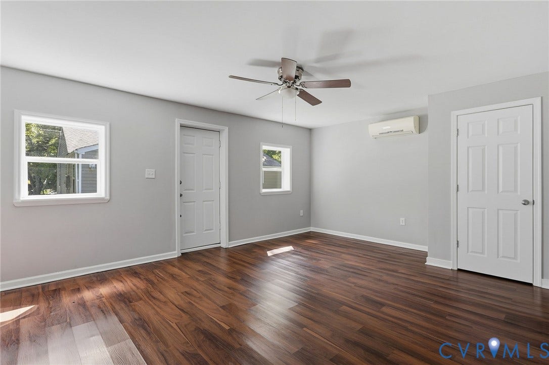 Wood-finish flooring, white paneled doors, ceiling fan with integrated lighting, two windows, and a wall-mounted climate control unit
