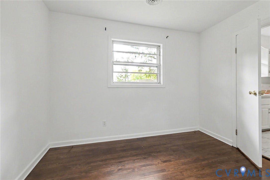 Dark wood-finish flooring with white baseboards, a single window with grids, and a white interior door with brass hardware