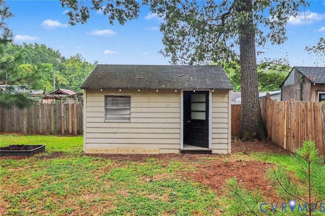 Detached storage shed with a solid door, surrounded by a wooden privacy fence, mature trees, and a raised garden bed