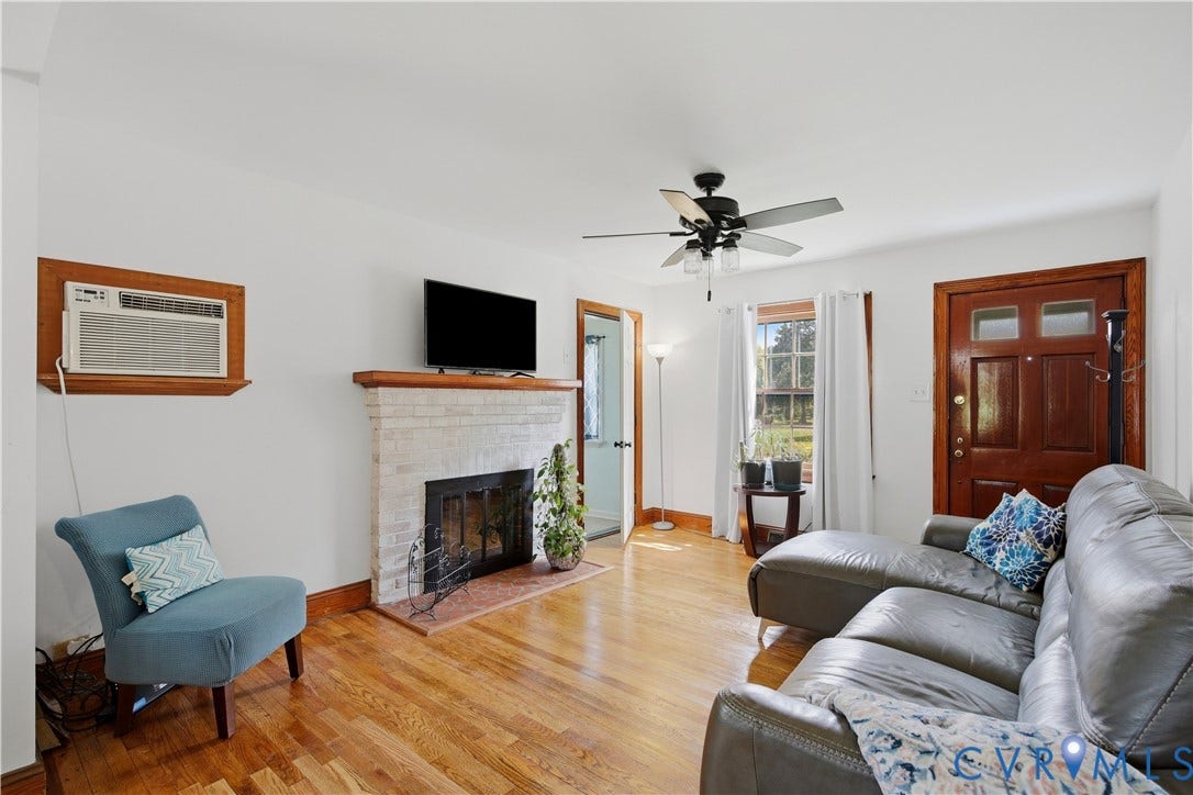 Hardwood flooring, white painted walls, a fireplace with a white brick surround and wood mantle, a ceiling fan, and a wood-paneled door