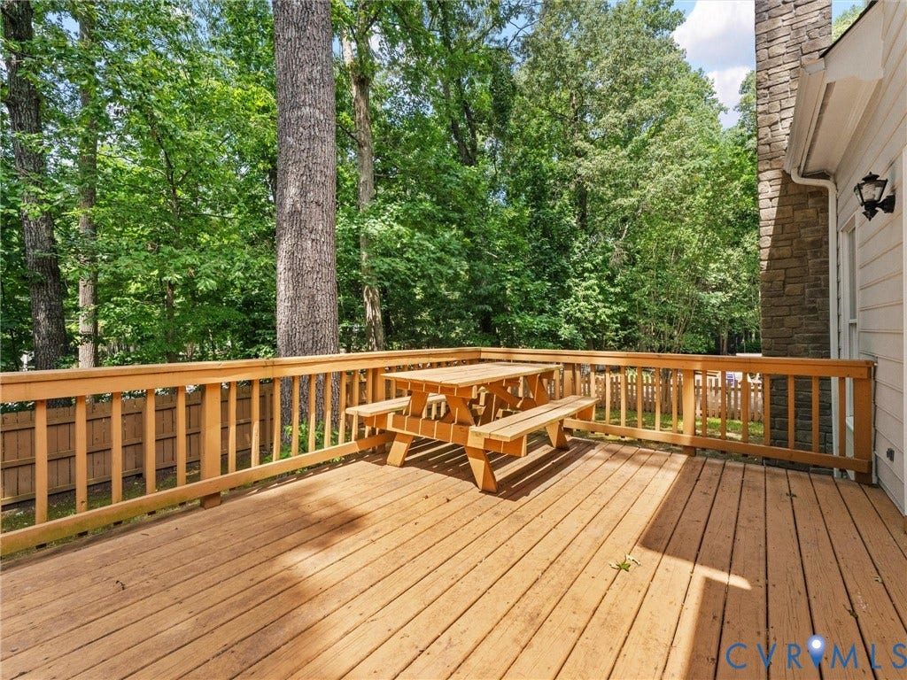 3700 Bremerton Drive, Wellesley, Henrico, VA 23233 Expansive wood deck with wood railings, surrounded by mature trees, featuring a stone-clad chimney and a wall-mounted exterior light fixture