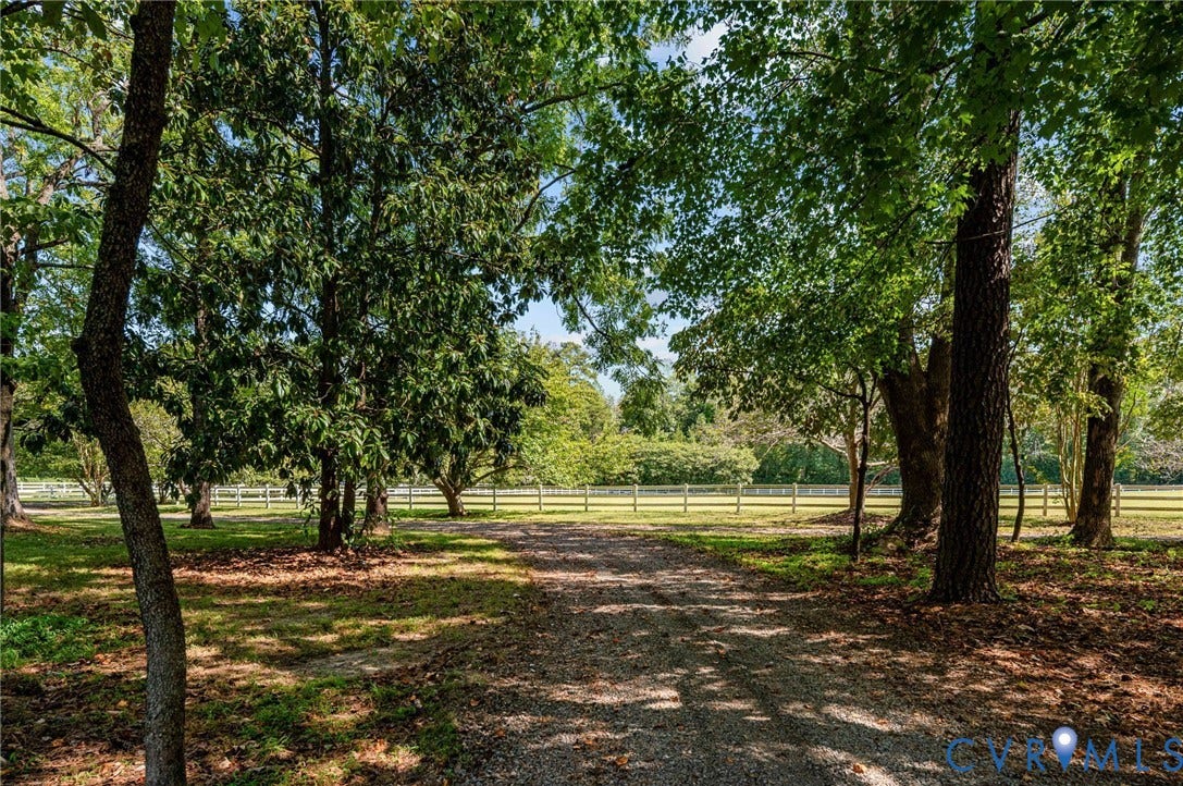 Gravel driveway framed by mature trees, leading to an expansive grassy area with a perimeter fence